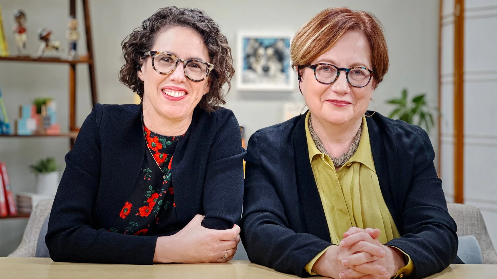 Two women seating close to each at a desk. They are wearing glasses and have a tender and positive look