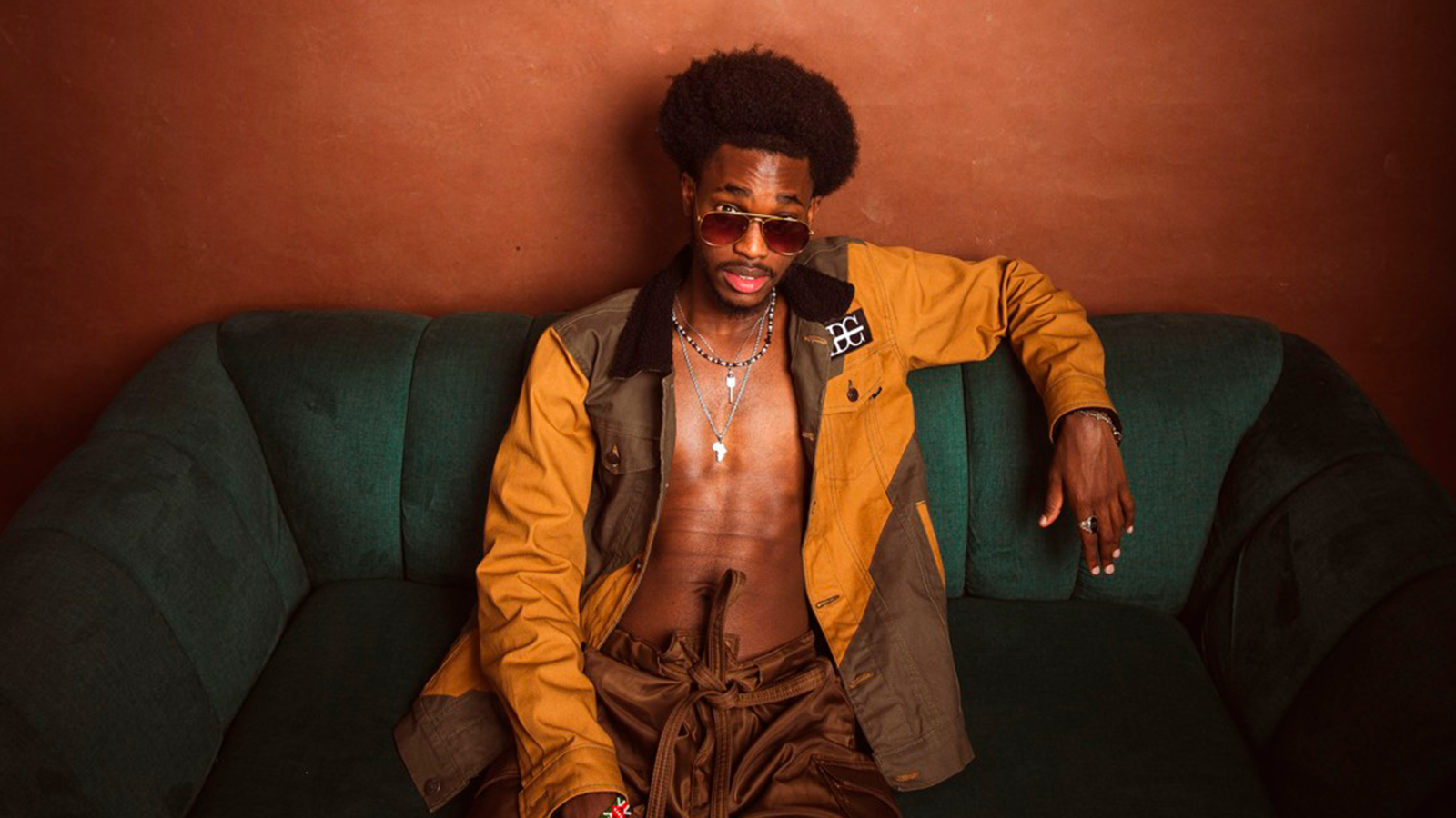 An artist sitting on a sofa wearing a yellow-brown jacket with no top in shades faces the camera