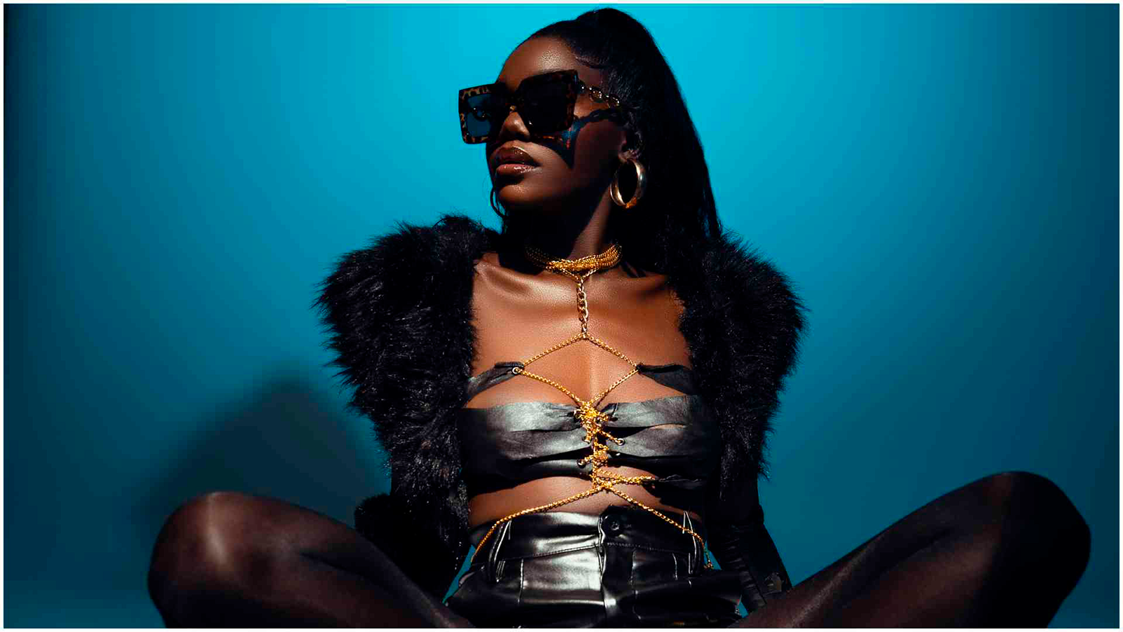 Female artist wears sunglasses looking to her right. She wears long dark hair and wears provocative outlook.