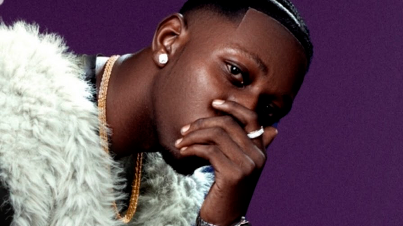 Closed up picture of an artist covering his mouth with one of his hands wearing a ring and a diamond ear ring and a white fur vest.