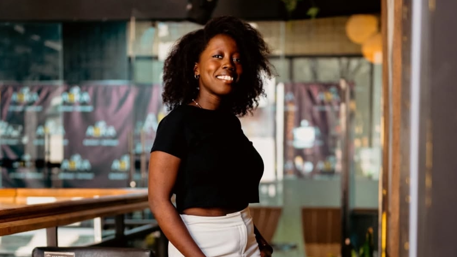 Young woman wearing a black top and white trousers stares at the camera at a cafe.
