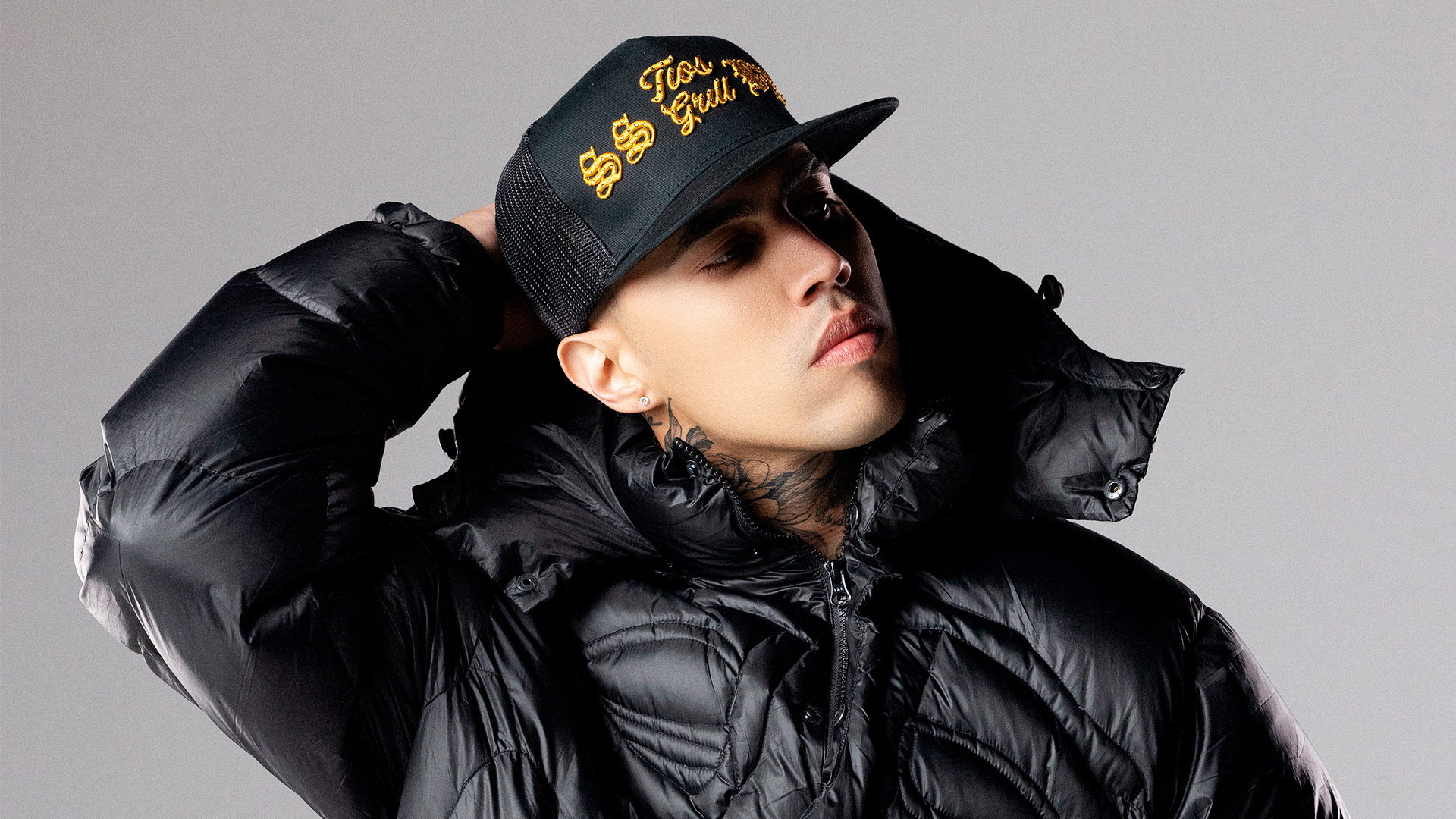 Singer Brytiago wears a black hoodie and a black cap looking to the side with challenging expression.