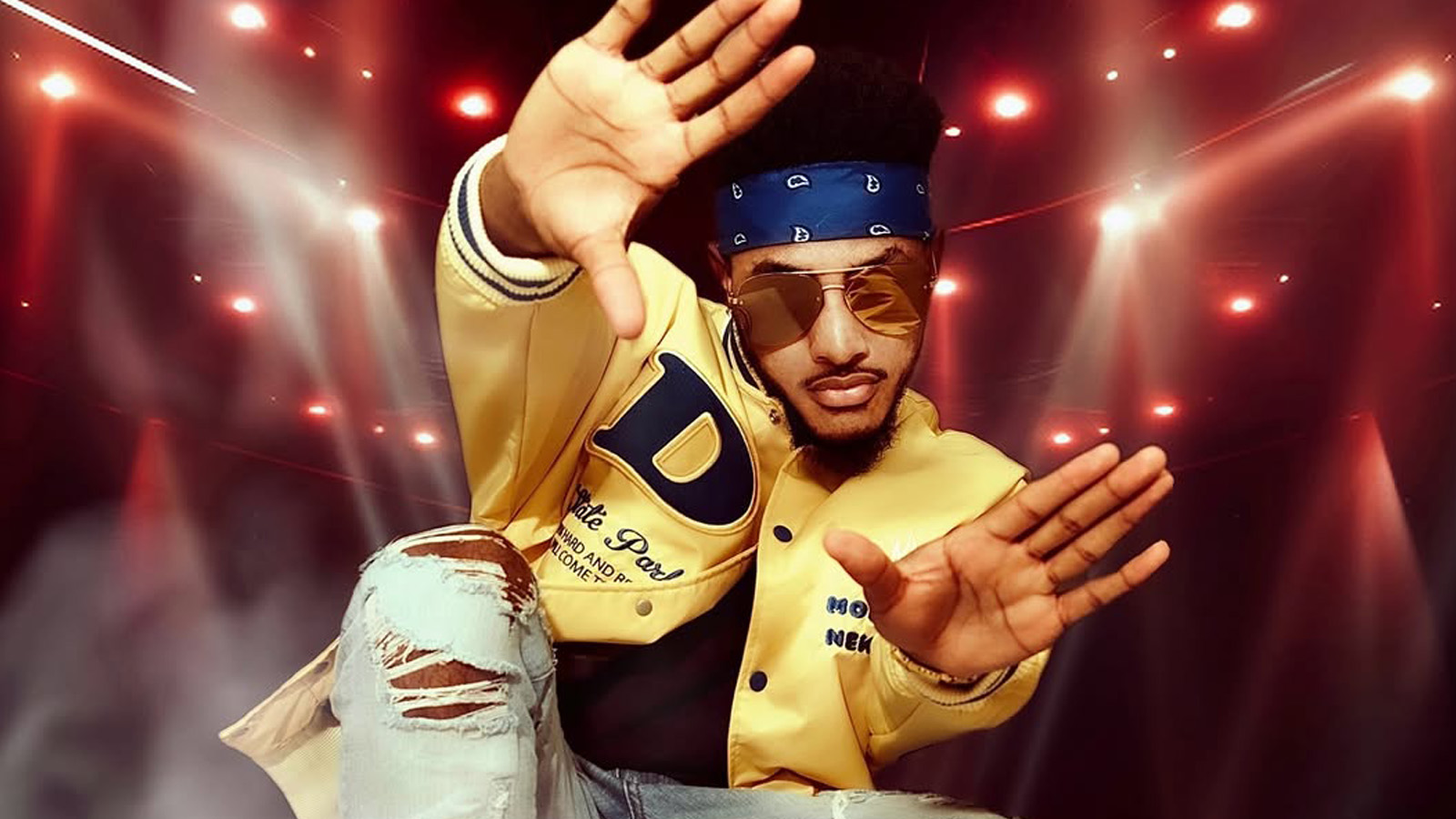 Haitian-Congolese artist Paska wearing sunglasses, a yellow bomber jacket and a headband showing their palms wide open to the camera